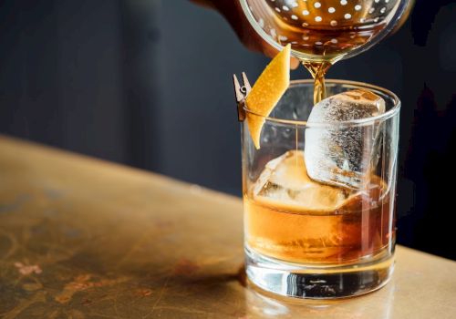 A glass of whiskey on the rocks with a cherry garnish, ice cube, and a citrus twist, poured from a decanter onto a wooden bar counter.