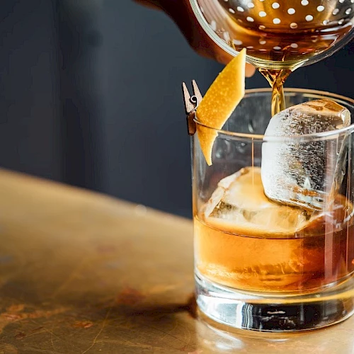 A glass of whiskey on the rocks with a cherry garnish, ice cube, and a citrus twist, poured from a decanter onto a wooden bar counter.