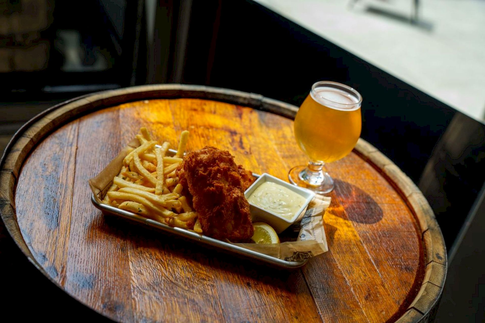 A round wooden table with a tray of fried chicken, french fries, a dipping sauce, and a glass of beer on the tray.