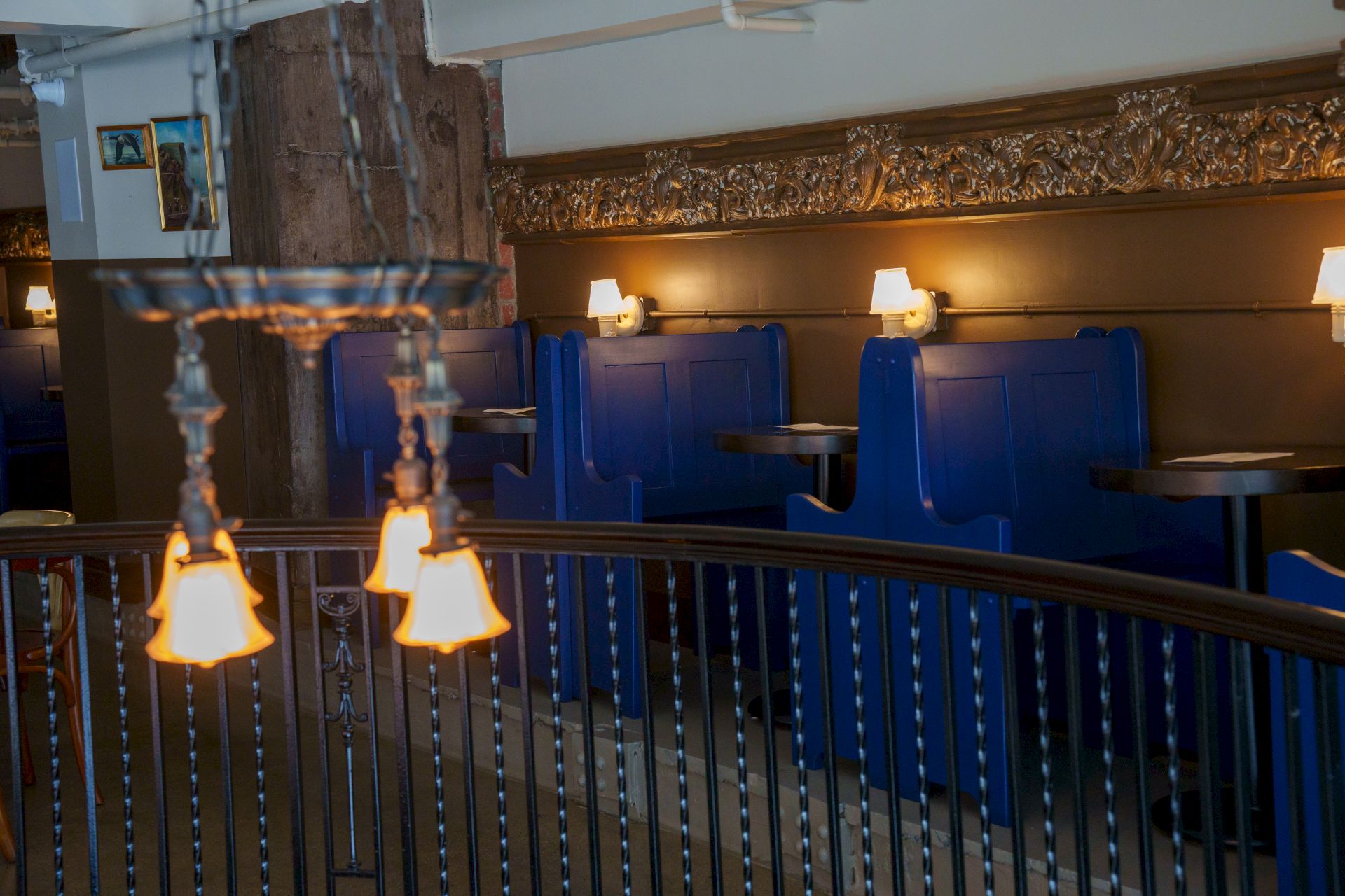 A cozy, dim restaurant with blue booths, small tables, warm wall lamps, and a decorative railing near the foreground.