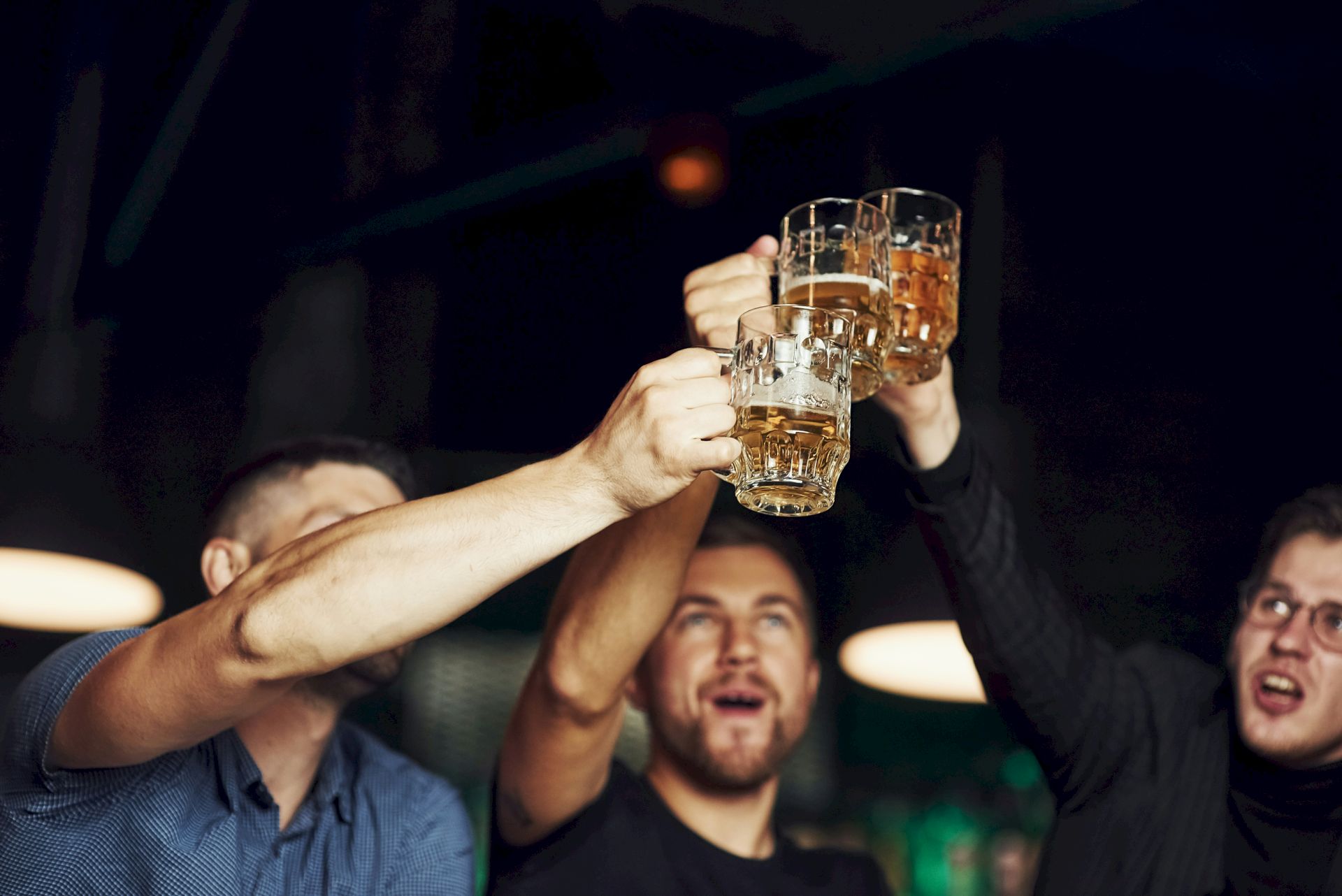 Friends clink beer glasses in a dim bar, toasting with amber drinks and cheering together in the glow.