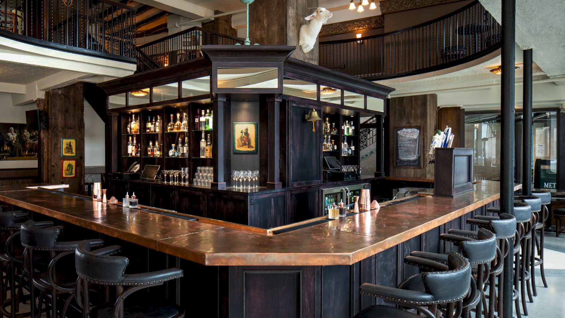 An ornate hotel bar with a stocked liquor shelf, copper/wood counters, and a hanging white dog decoration above the staircase.